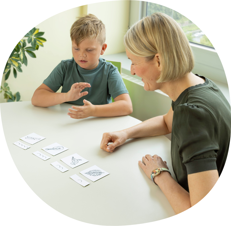 Ein Therapiekind sitzt mit seiner Lerntherapeutin an einem Tisch. Vor ihm liegen Zeichnungen von Objekten. Es hat die passenden Silbenbögen zu den jeweiligen Objektzeichnungen zugeordnet. Es klatscht die Silben während es die Wörter liest. Seine Lerntherapeutin sitzt daneben und ermutigt das Therapiekind.