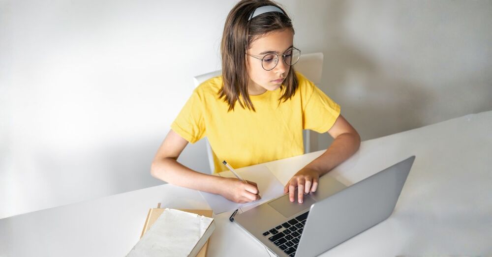 Ein junges Teenager-Mädchen sitzt konzentriert vor einem Laptop an einem Tisch. Vor ihm liegt ein leeres Blatt Papier. In der rechten Hand hält es einen Stift. Es setzt zum Schreiben an. Daneben liegt ein Bücherstapel auf dem Tisch.