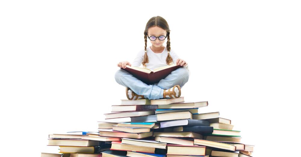 Ein Mädchen sitzt im Schneidersitz auf einem großen Bücherhaufen und liest ein großes Buch.