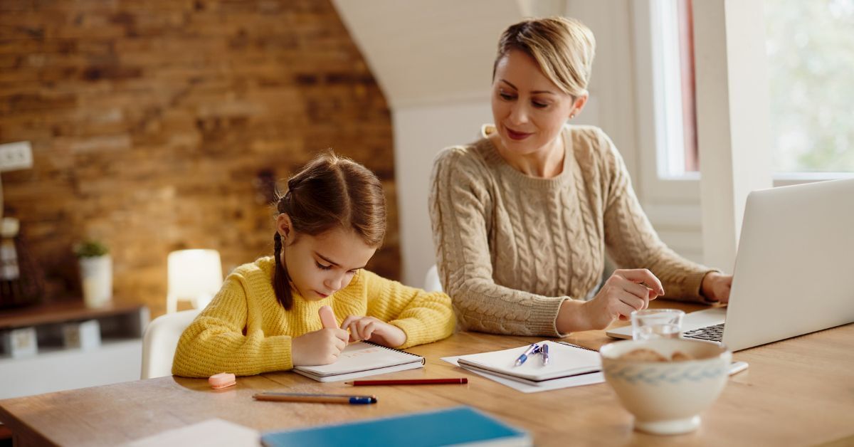 Ein kleines Mädchen radiert etwas in ihrem Notizblock, während ihre Mutter daneben zu Hause am Laptop arbeitet und zu ihrer Tochter rüber schaut.